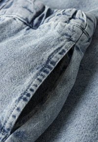 Svetlo modro jeansa tkanina z teksturirano površino, ki vključuje naborke na šivih in poševni žep. Podrobnosti prikazujejo šive in raztrgane robove.