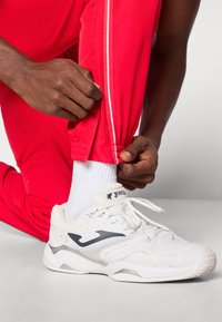 Bele športne copate z sivimi detajli in teksturirano podplatom, skupaj s svetlo rdečimi tekaškimi hlačami, ki imajo beli stranski trak.