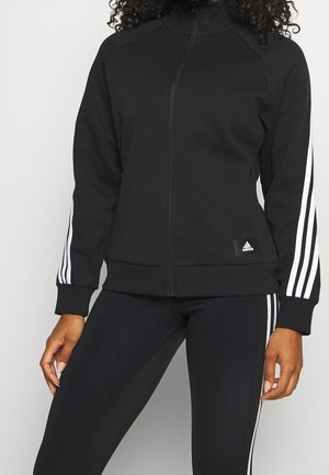 Vrouw draagt een zwarte Adidas ritsjack en bijpassende legging met witte drie-streep details op de mouwen en zijkanten.