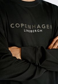 Camisa negra de manga larga con "COPENHAGEN" impreso en letras gris oscuro y "LINDBERGH" en blanco. Tejido suave con un corte relajado.