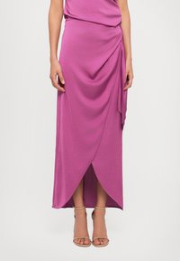 Roze wikkelrok van zachte stof, met draperingsdetails, een voorloop en gecombineerd met lichtgekleurde sandaaltjes.