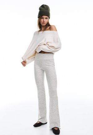 Lichtbeige oversized trui met off-shoulder ontwerp, gecombineerd met lichtgrijze uitlopende broek en bruine platte schoenen. Gestructureerde gebreide muts, olijfgroen.