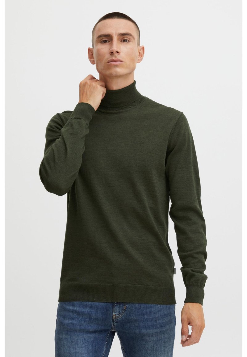 Hombre vestido con un suéter de cuello alto verde oscuro y jeans azules, ajustando el cuello con una mano frente a un fondo claro y uniforme.