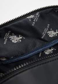 Černý vnitřek tašky s hladkou texturou, zdobený opakujícím se bílým logem U.S. Polo Assn. a nápisem "Since 1890".