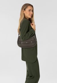 Sac à main en cuir gris au design incurvé, sécurisé par des accents en métal argenté. Porté à l'épaule par un mannequin vêtu d'un costume vert.