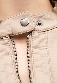 Κοντινό πλάνο ενός μπεζ δερμάτινου μπουφάν με μερικώς ανοιγμένο φερμουάρ και μεταλλικό κουμπί τύπου τρουκ με την επιγραφή «ONLY» κοντά στον γιακά.