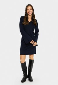Donna sorridente che indossa un vestito lungo blu navy con maniche a campana e stivali neri fino al ginocchio, in piedi contro uno sfondo semplice.