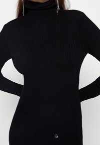 Černé žebrované šaty s rolákem a dlouhými rukávy. Mají přiléhavý střih a kruhový detail zlatého tónu na spodním lemu.