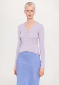 Lichtpaarse langemouwshirt met een V-hals en vijf knoppen aan de voorkant, gecombineerd met een lichtblauwe rok. Glad, aansluitend materiaal.