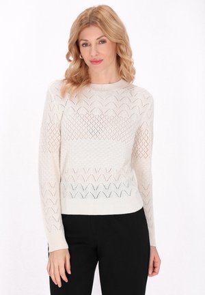 Pulover tricotat bej cu modele în diamant și zigzag, mâneci lungi și decolteu rotund, asortat cu pantaloni negri.