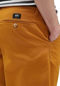 Man med senapsgula Vans-byxor, med handen i framfickan, visar knappen på bakfickan och Vans-logotypens patch.