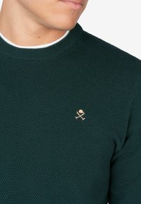 Ciemnozielony sweter o fakturze z prążkowanym kołnierzem, z małym haftowanym jasnoróżowym logo czaszek i róż na lewej piersi.
