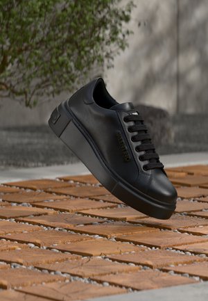 Čierna kožená Valentino teniska s hrubou podrážkou a čiernymi šnúrkami vznášajúca sa nad vonkajším povrchom z drevených dlaždíc a kamienkov.