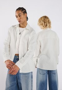 Chemise blanche à manches longues texturée avec un bouton sur le devant, portée sur un haut sans manches, associée à un jean bleu ample.