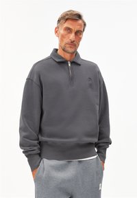 Grijze rits-trui met een hoge kraag, textuurstof en een geperst letterontwerp op de borst. Draagt met grijze joggingsbroek.