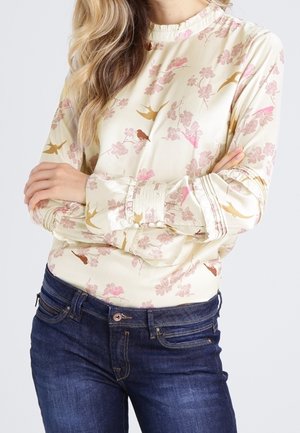Blusa crema con motivi floreali rosa e uccelli dorati, in morbido materiale di seta, con collo alto e polsini abbottonati, abbinata a jeans blu scuro.