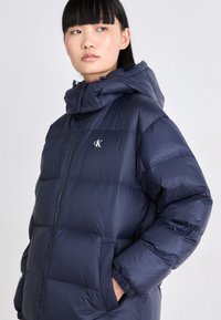 Mörkblå puffjacka med dragkedja, elastiska ärmslut och huva. Har en diskret logotyp på bröstet; tillverkad av ett glänsande, isolerat material.