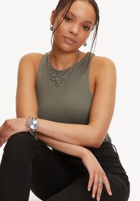 Silberne Uhr und Schmuck auf einem grünen Tanktop. Die Uhr hat ein rundes Ziffernblatt und ein dickes Gliederarmband. Schlichte silberne Ohrringe und ein Ring.