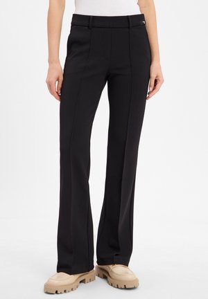 Femme portant un pantalon noir droit sur mesure et des chaussures beige à semelles épaisses, debout devant un fond blanc.