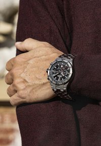 Orologio in acciaio inossidabile argentato con un quadrante nero, più quadranti e numeri audaci. Indossato su un polso contro uno sfondo di tessuto testurizzato color bordeaux.