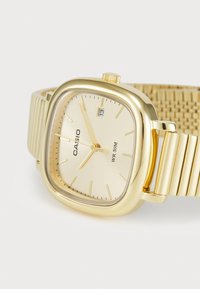 Montre Casio dorée avec un cadran carré, un cadran beige texturé, des aiguilles fines et un affichage de date à 3 heures ; bracelet en métal intégré.