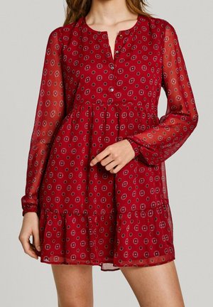 Kobieta ubrana w czerwoną sukienkę z długim rękawem, sięgającą kolan, z okrągłymi wzorami i zapięciem na guziki przy dekolcie, pozująca na jednolitym tle.