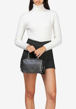 Sac à main noir texturé avec quincaillerie dorée, forme arrondie et double poignées supérieures, tenu par une personne portant un pull à col roulé côtelé blanc et une jupe rayée.