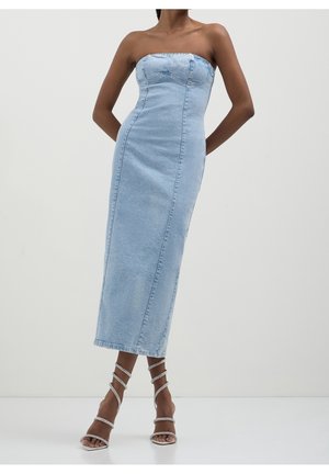 Frau trägt ein hellblaues trägerloses Denim-Midikleid mit tailliertem Oberteil und weißen, hochhackigen Sandalen mit Riemchen, steht auf einem weißen Boden.