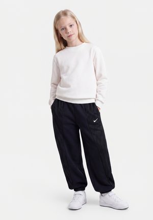 Jeune fille aux longs cheveux blonds portant un sweat-shirt blanc, un pantalon de jogging Nike noir et des baskets blanches, debout les mains dans les poches.