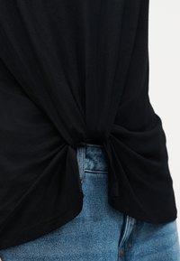 Μαύρο υφασμάτινο τοπ δεμένο με κόμπο στη μέση πάνω από μπλε τζιν, αναδεικνύοντας λεπτομέρεια casual εμφάνισης.