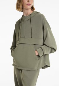 Zelena oversized hoodie s kapuljačom na šnuru, prednjim džepom i teksturiranim materijalom. Ima široke rukave i skraćeni kroj.