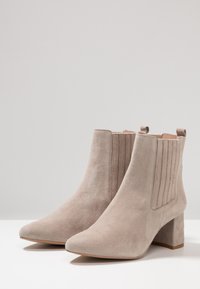 Botas de tobillo de ante en un color beige claro con punteras afiladas, paneles laterales texturizados y tacones de bloque. Acabado suave con un detalle mínimo.