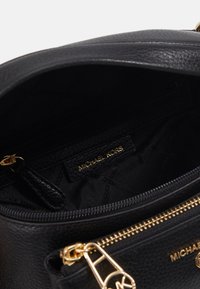 Wnętrze czarnej skórzanej torebki pokazujące kieszeń na zamek, złote akcenty i metkę Michael Kors. Teksturowana powierzchnia z materiałowym wykończeniem.
