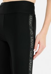 Leggings atletici neri con una texture liscia, caratterizzati da un pannello laterale decorato con un dettaglio logo riflettente. Design con vita alta.