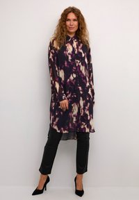 Tunica lunga con bottoni in un motivo astratto viola scuro e crema, realizzata in tessuto leggero. Abbinata a pantaloni di pelle nera e tacchi.