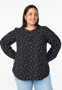 Blusa nera a fantasia con scollatura rotonda, maniche lunghe, chiusura a bottone e una texture morbida con disegni geometrici bianchi.