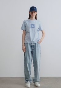 Γαλάζιο βαμβακερό μπλουζάκι με τετράγωνο λογότυπο CK, συνδυασμένο με φαρδιά γαλάζια τζιν και λευκά αθλητικά παπούτσια. Φοράει σκούφο ναυτικού.