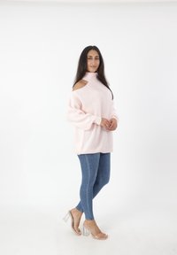 Donna in sandali trasparenti con tacco, jeans attillati blu e un maglione rosa chiaro lavorato a maglia con dettagli spalla scollata, su sfondo bianco.