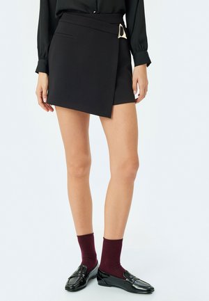 Falda negra envolvente con detalle de hebilla lateral, textura suave y un ajuste entallado. Combinada con calcetines burdeos y mocasines negros brillantes.