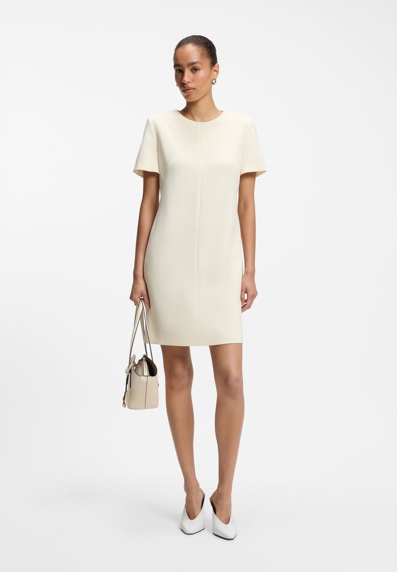 Femme portant une robe crème à manches courtes, longueur genou, tenant un sac à main beige et portant des escarpins blancs à bout pointu sur un fond uni.