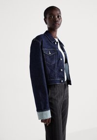 Dark blue denim jacket with gold stitching, two chest pockets, and contrasting light blue cuffs. Worn over a white shirt and pinstriped pants.