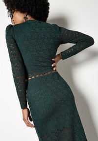 Top de manga larga verde con encaje y dobladillo festoneado, combinado con una falda a juego. Patrón floral visible a través de una tela semi-translúcida.