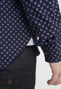 Camicia blu navy con motivo a goccia bianca, maniche lunghe e polsini con bottoni. Indossata sopra un denim scuro, con un logo sottile sull'orlo. Tessuto dalla texture liscia.
