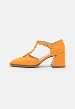 Chaussure orange en cuir à bout fermé avec bride en T, talon bloc moyen et fermeture réglable par boucle sur fond blanc.