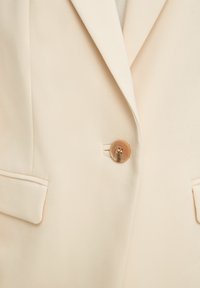 Blazer beige clair avec un seul bouton marron, col à encoche, et deux poches avant à rabat visibles sur la texture du tissu.