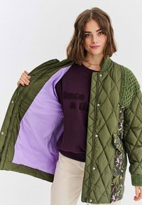 Prošívaná olivově zelená bunda s fialovou podšívkou a květinovým vzorem na zádech. Má žebrované pletené detaily a zapínání na druky.