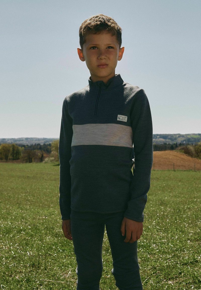 Garçon aux cheveux courts et bouclés portant un t-shirt à manches longues rayé gris foncé et gris clair, debout sur de l'herbe verte avec des arbres et des collines en arrière-plan.