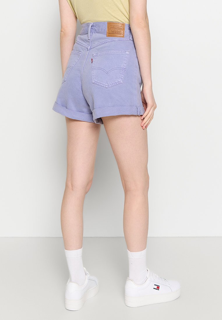 Lichtpaarse spijkershorts met opgerolde zomen, achterzakken en een leren bruine patch aan de tailleband; gecombineerd met witte sneakers.