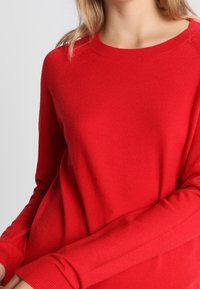 Rød strikket sweater med lange ærmer, ribbede manchetter og rund halsudskæring. Blød tekstur, afslappet pasform og minimal sømdetaljering.