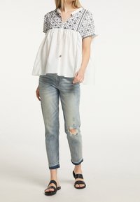 Haut à manches courtes blanc et à motifs avec une silhouette froncée, associé à un jean bleu clair déchiré et des sandales noires à enfiler.